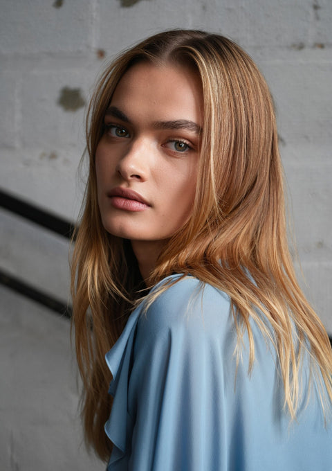 AI Fashion-Woman with long blonde hair wearing a light blue top against a textured wall.